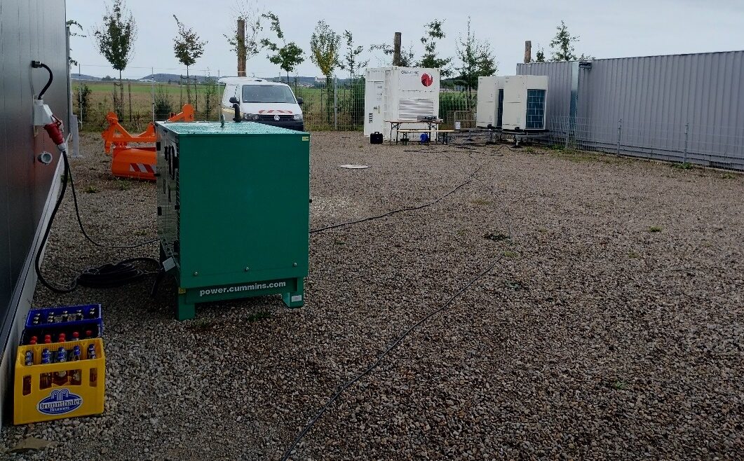 Grünes Notstromaggregat auf einem Schotterplatz neben Containern